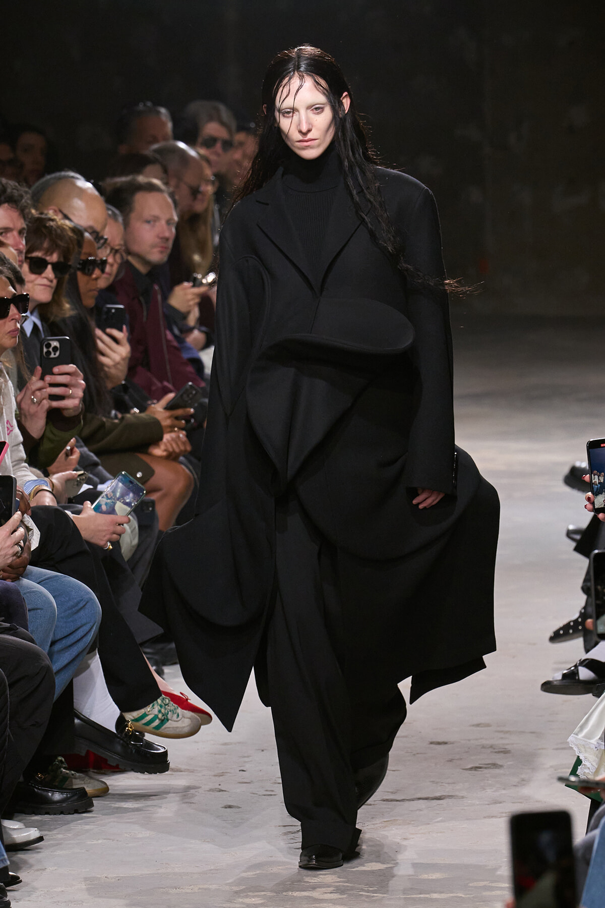 Model walks a runway in an oversized black sculptural coat as the audience watches and photographs her.