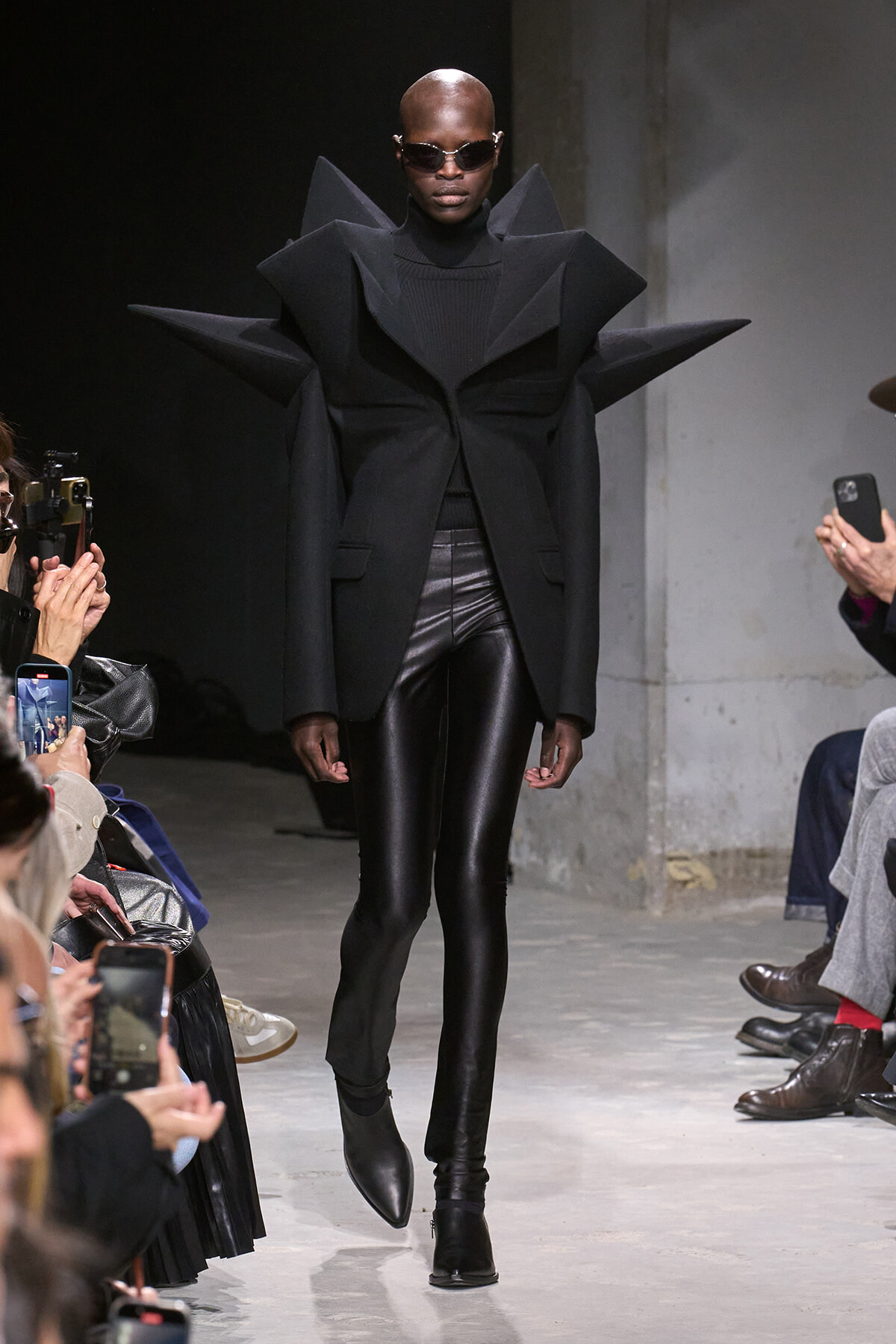 Model on a runway wearing a dramatic black sculptural coat with oversized angular shoulders and dark sunglasses.