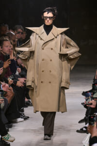 Model on a runway wearing a dramatic beige trench with oversized angular shoulders and cape-like sleeves.
