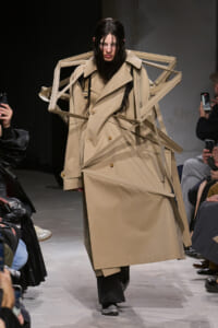 Model on a runway wearing a beige oversized trench coat with architectural strap details and bold silhouette, audience photographing the look nearby.