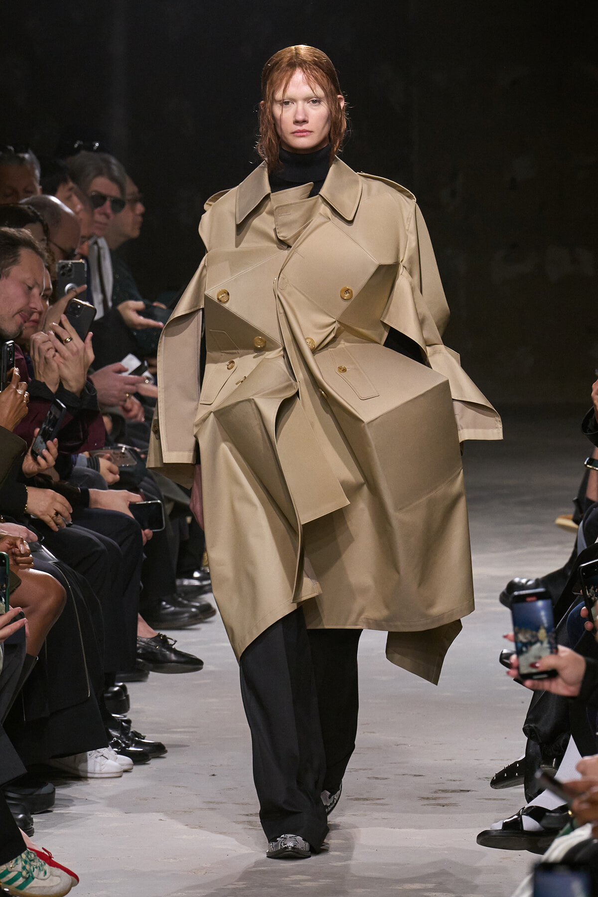 Model walks the runway in a dramatic beige trench coat with sculpted panels and large lapels, audience watching on both sides