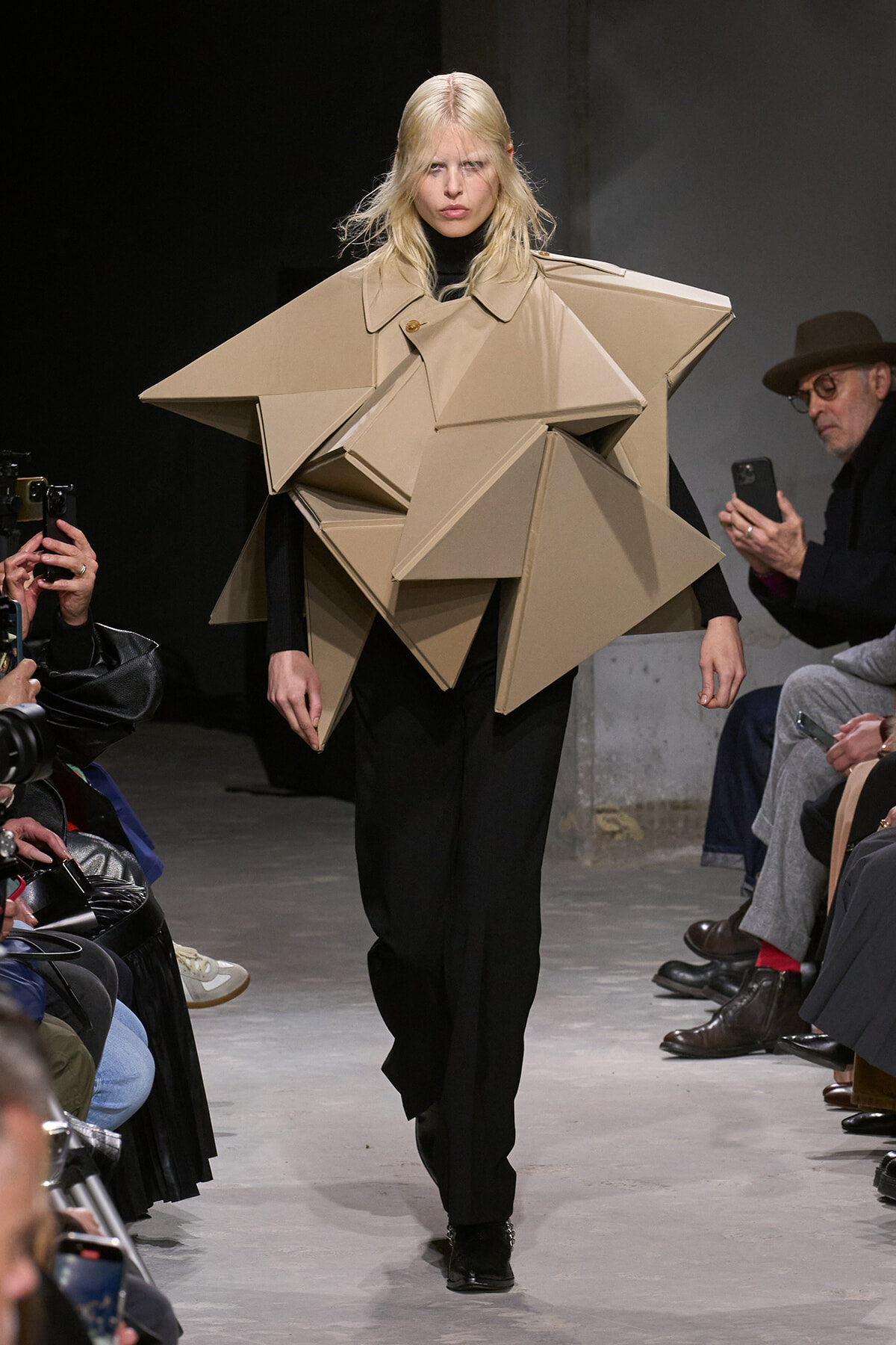Model walks a runway in a large beige angular coat made of geometric shapes for an avant-garde fashion show.