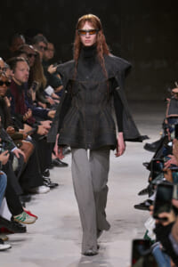 Runway model in a dark denim cape-style coat with orange stitching over a black turtleneck and gray wide-leg pants, wearing sunglasses.