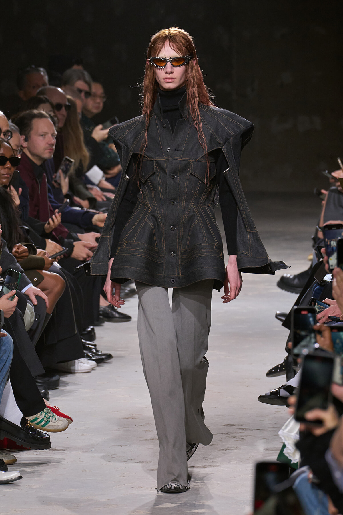 Runway model in a dark denim cape-style coat with orange stitching over a black turtleneck and gray wide-leg pants, wearing sunglasses.