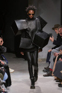 Model walks the runway in a black, geometric coat with oversized cube shapes and matching black leather pants, finished with spiked boots and sunglasses on a catwalk.