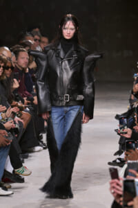 Model on a runway wearing an oversized black leather jacket with exaggerated shoulders, a black turtleneck, belt, blue jeans, and long black fur pants; audience watches.