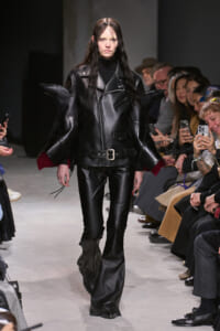 Model walking a runway in an avant-garde black leather outfit with oversized sculpted shoulders and matching pants, audience watching nearby.