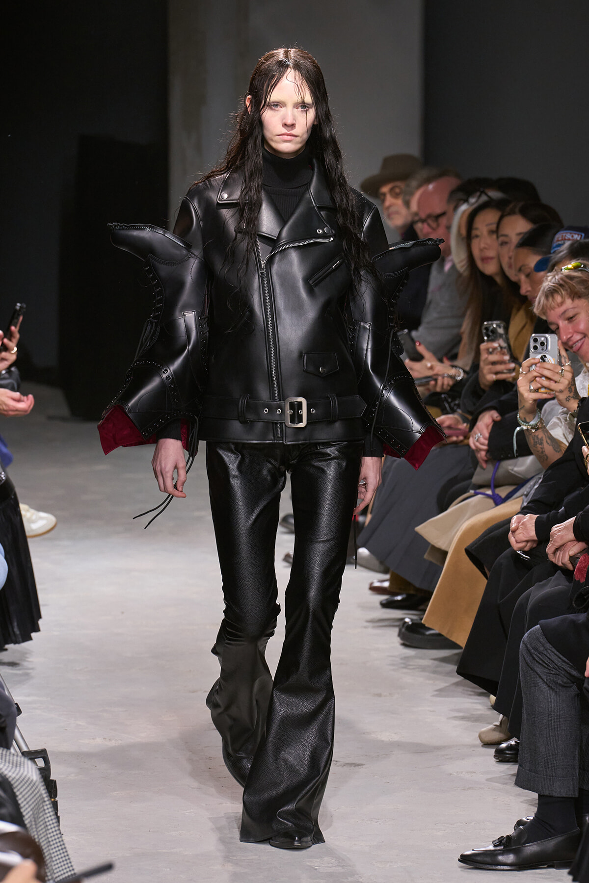 Model walking a runway in an avant-garde black leather outfit with oversized sculpted shoulders and matching pants, audience watching nearby.
