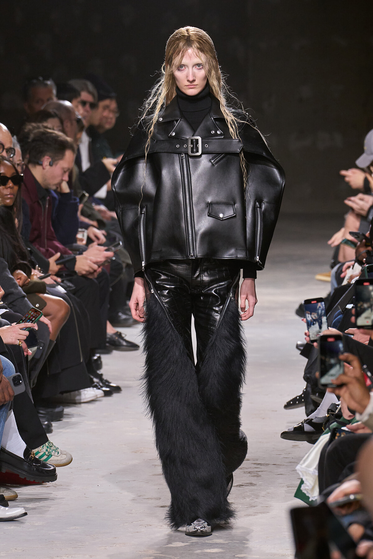 Model walks a runway in an oversized black leather jacket with a wide belt, paired with glossy black pants and long furry leg coverings.