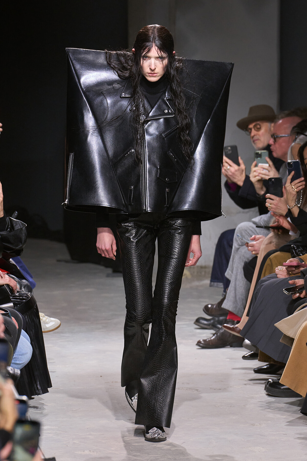 Model walks the runway in an oversized black leather angular jacket and snakeskin pants.