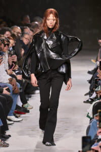 Model walks the runway in an oversized black leather jacket with wide sleeves and black trousers, audience watching closely.