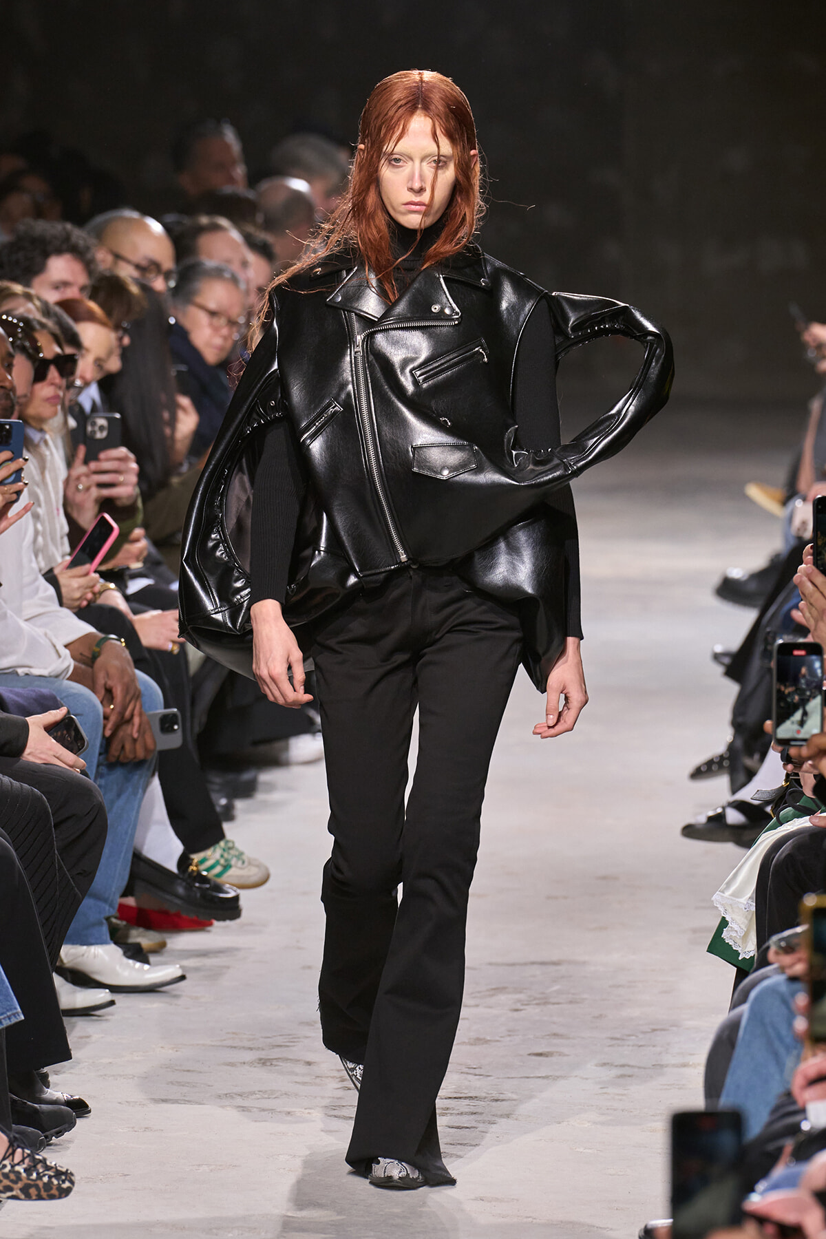 Model walks the runway in an oversized black leather jacket with wide sleeves and black trousers, audience watching closely.