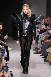 Female model walks the runway in a black patent leather coat with oversized spiked shoulders and a waist belt.