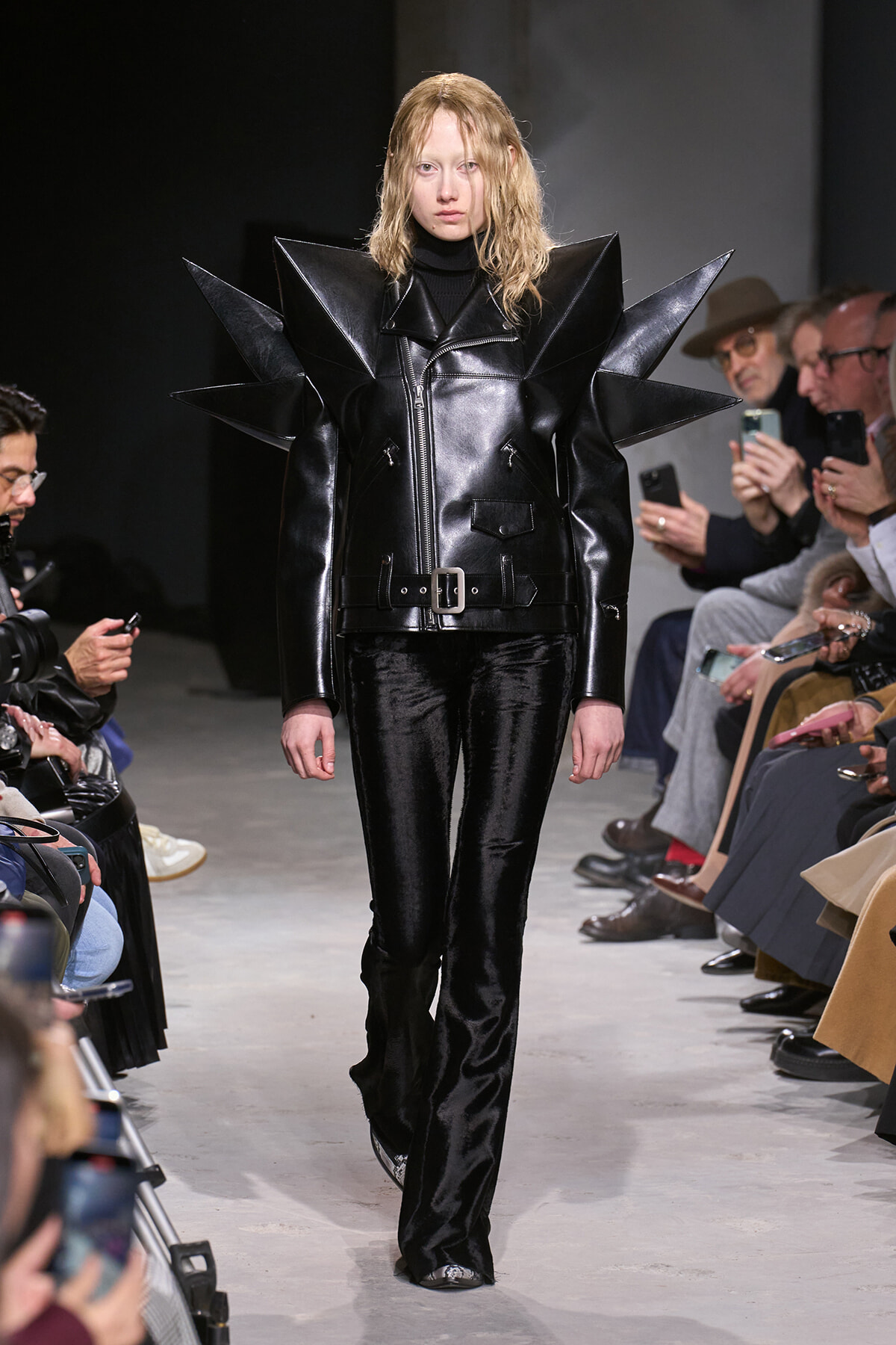 Female model walks the runway in a black patent leather coat with oversized spiked shoulders and a waist belt.
