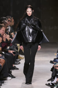 Model walks the runway in a glossy black oversized leather cape jacket with a waist belt, paired with black pants.