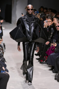 Model walking a runway in an all-black avant-garde leather outfit with oversized angular shoulders, sunglasses, and pointed-toe boots.