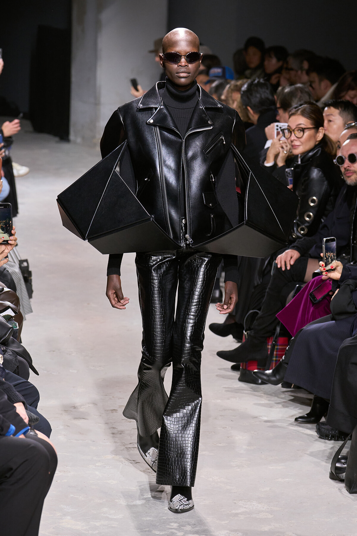Model walking a runway in an all-black avant-garde leather outfit with oversized angular shoulders, sunglasses, and pointed-toe boots.