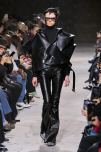 Model walks a fashion show wearing an oversized black leather geometric cape over a black turtleneck and pants, sunglasses on.