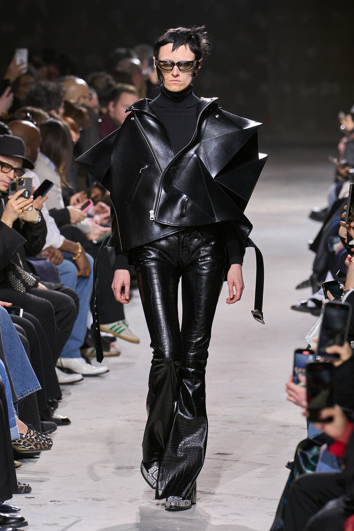 Model walks a fashion show wearing an oversized black leather geometric cape over a black turtleneck and pants, sunglasses on.