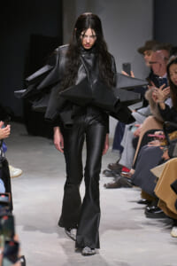 Model on a runway wearing an oversized black leather ensemble with large angular, pyramid-shaped shoulder pieces; audience watches.
