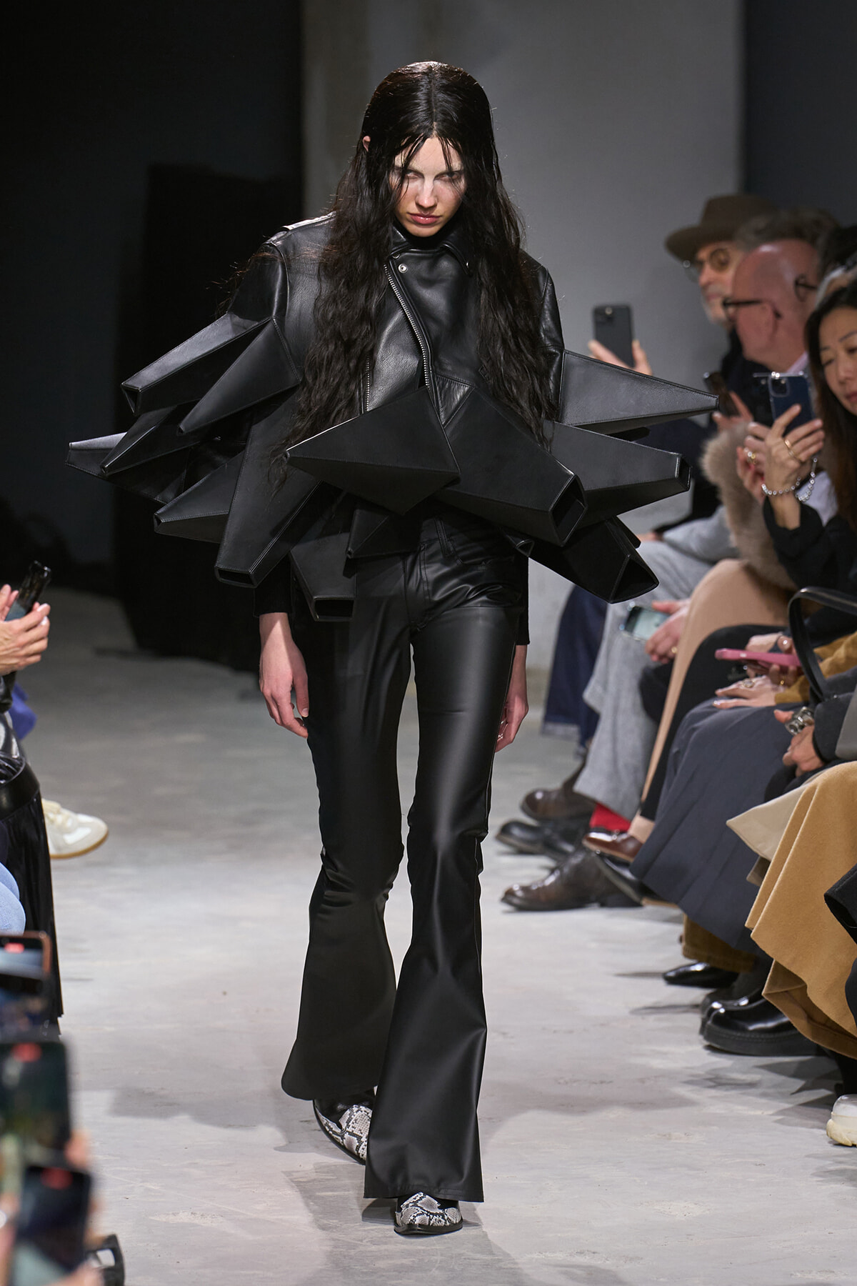 Model on a runway wearing an oversized black leather ensemble with large angular, pyramid-shaped shoulder pieces; audience watches.