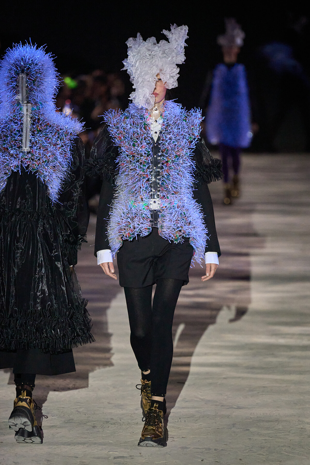 Runway model in a blue spiky embellished jacket over a white shirt, black shorts, black tights, and gold boots, with a large white floral headpiece.