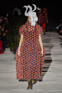 Model on a runway wearing a vibrant multicolor crochet gown with oversized sleeves and a sculptural white headpiece.