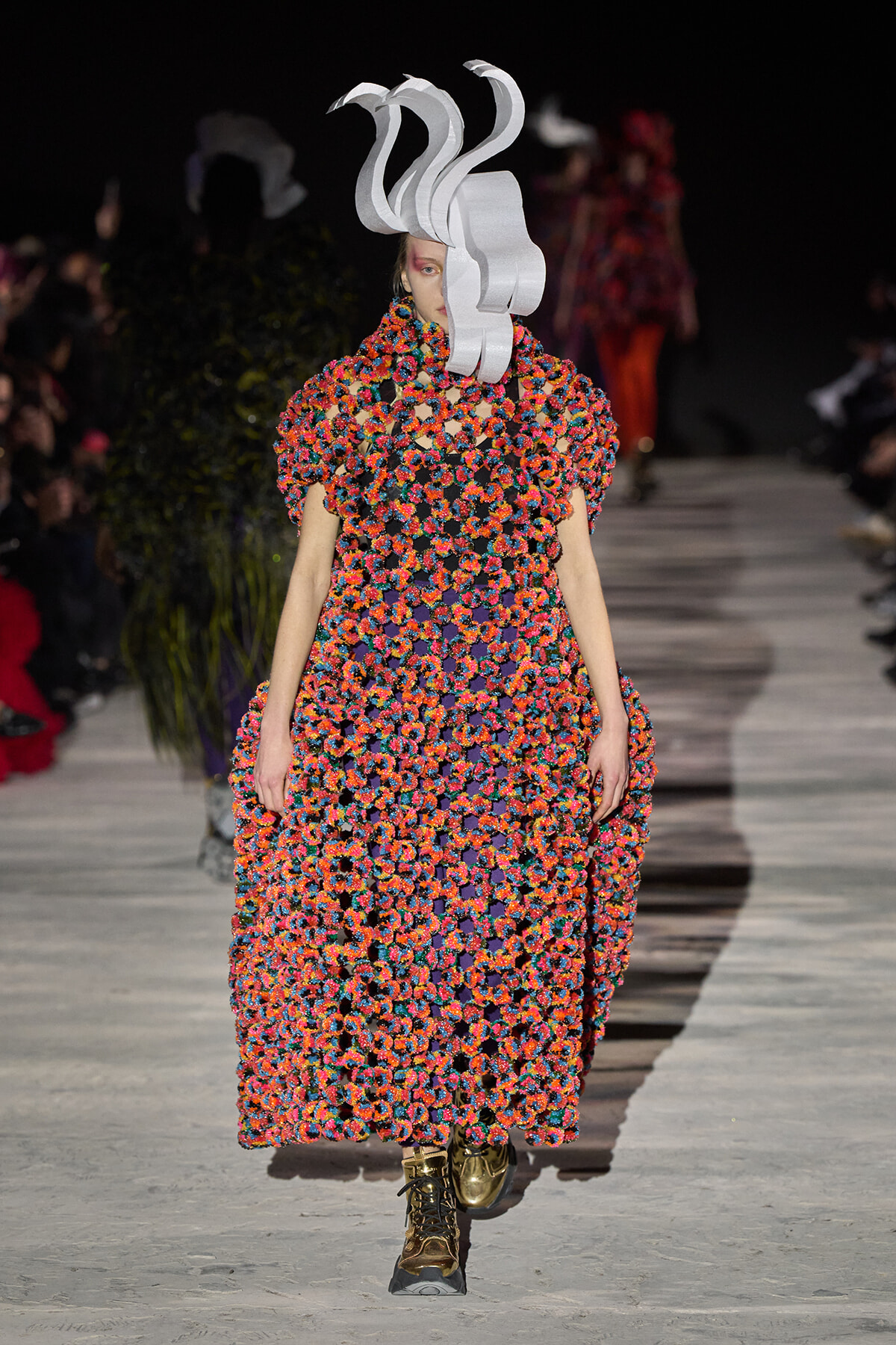 Model on a runway wearing a vibrant multicolor crochet gown with oversized sleeves and a sculptural white headpiece.