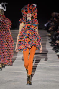 Model on a runway wearing a bright multicolor textured floral outfit with orange tights and metallic boots.