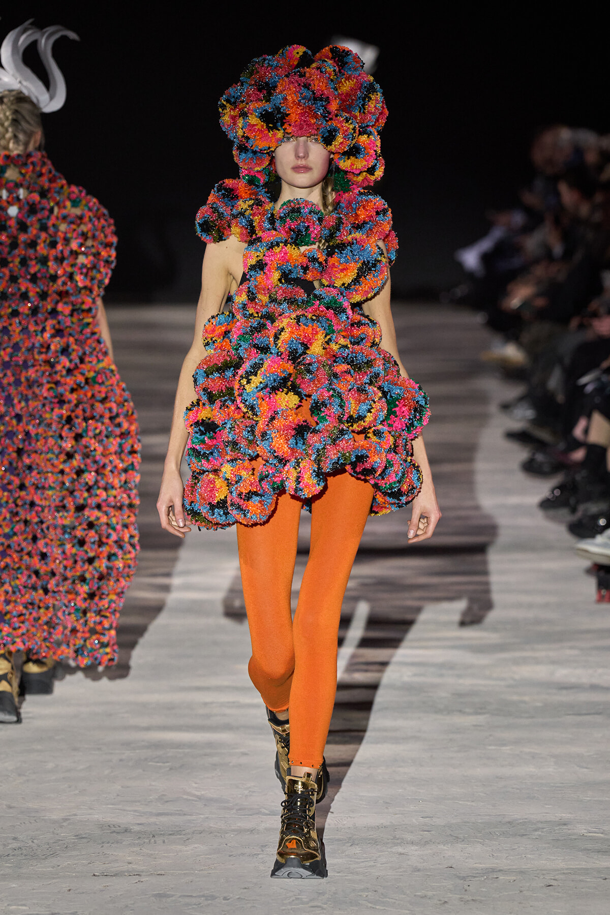Model on a runway wearing a bright multicolor textured floral outfit with orange tights and metallic boots.