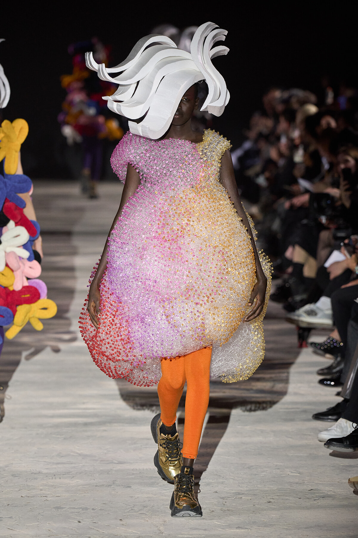 Model walks a fashion runway in a pink-to-orange beaded bubble dress with an oversized white sculptural headpiece; orange tights and gold boots visible.