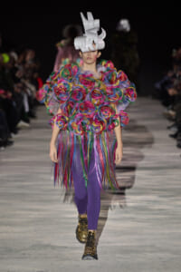 Model walks the runway in a vivid, multicolored ruffle top with a long fringed skirt and purple tights, wearing a sculptural white headpiece and metallic boots.