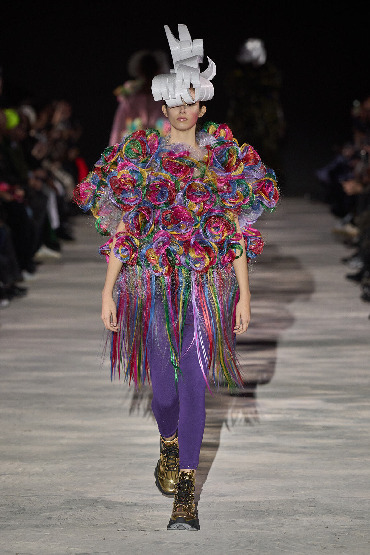 Model walks the runway in a vivid, multicolored ruffle top with a long fringed skirt and purple tights, wearing a sculptural white headpiece and metallic boots.