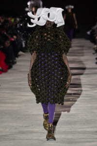 Model on a fashion runway in a black crocheted dress with purple lining, and a large white sculptural hat with gold boots.