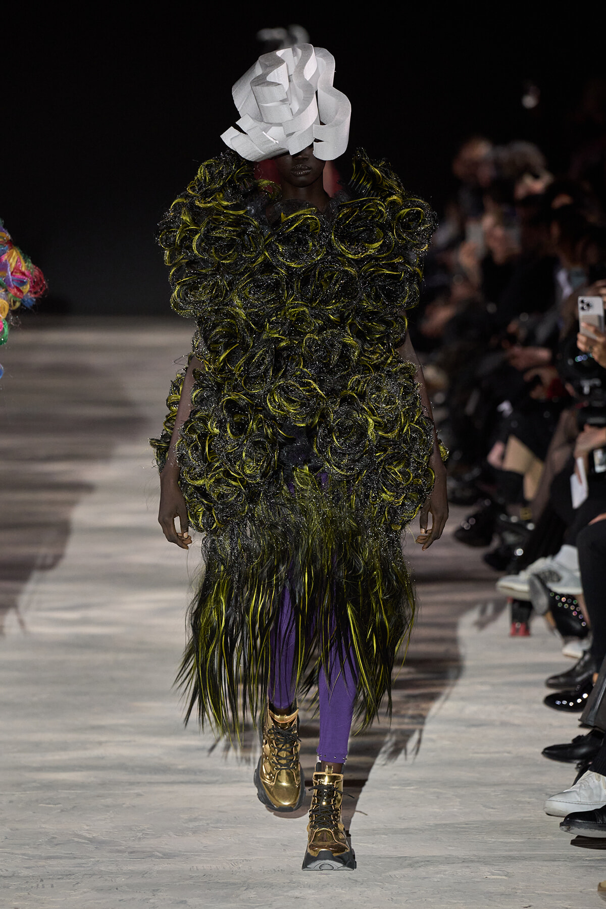Runway model wearing a dramatic black-green textured coat with purple pants, gold boots, and a large white sculptural headpiece.