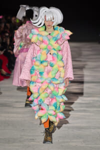 Model walks the runway in a pink quilted coat adorned with multicolor fluffy pom-poms and a large white sculptural hat.