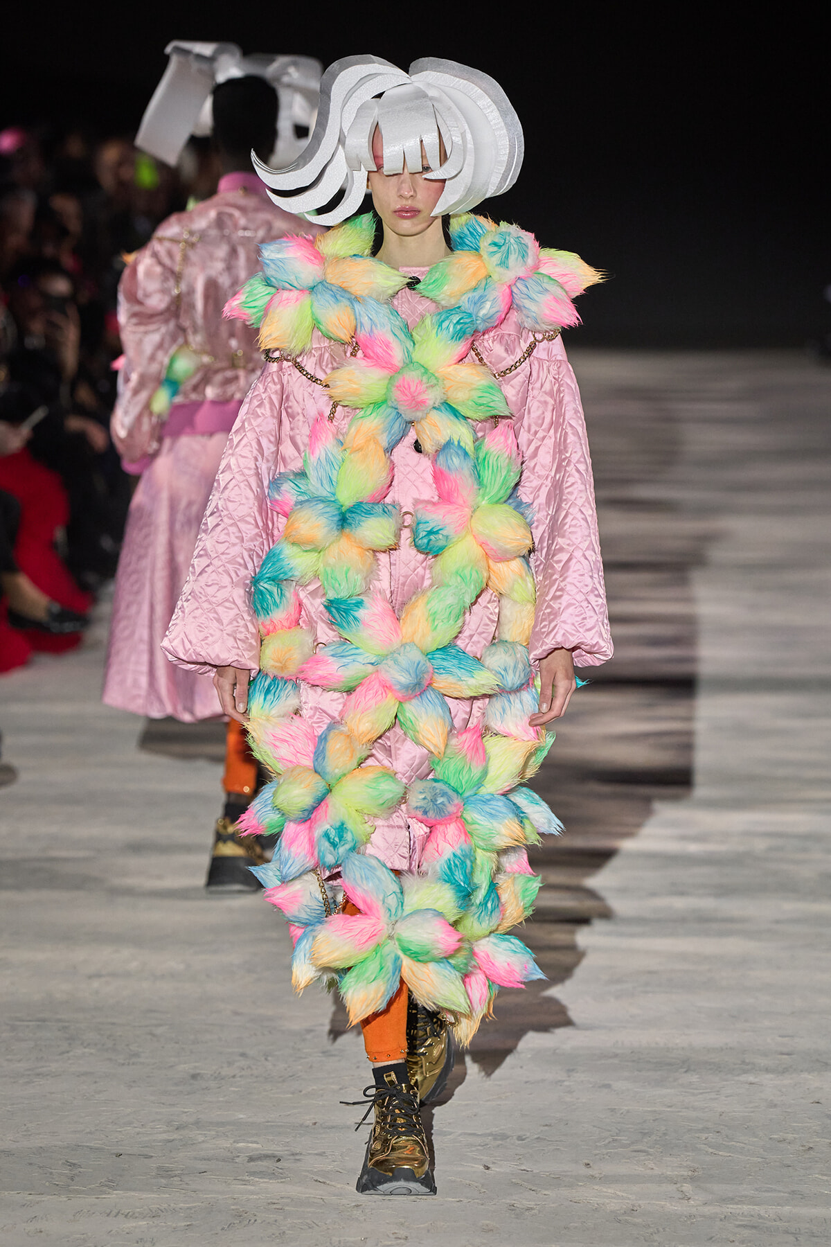 Model walks the runway in a pink quilted coat adorned with multicolor fluffy pom-poms and a large white sculptural hat.