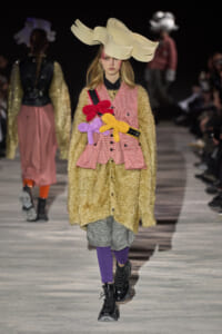 Model on runway wearing an oversized cream sculptural hat, gold textured coat, pink check vest with colorful plush flowers, plaid shorts, purple tights, and black boots.