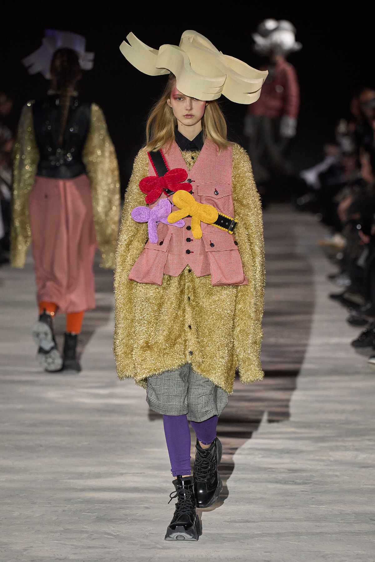 Model on runway wearing an oversized cream sculptural hat, gold textured coat, pink check vest with colorful plush flowers, plaid shorts, purple tights, and black boots.