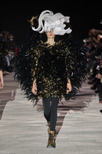 Runway model in a velvety black feathered cape with gold accents and a large white sculptural headpiece, walking the runway.