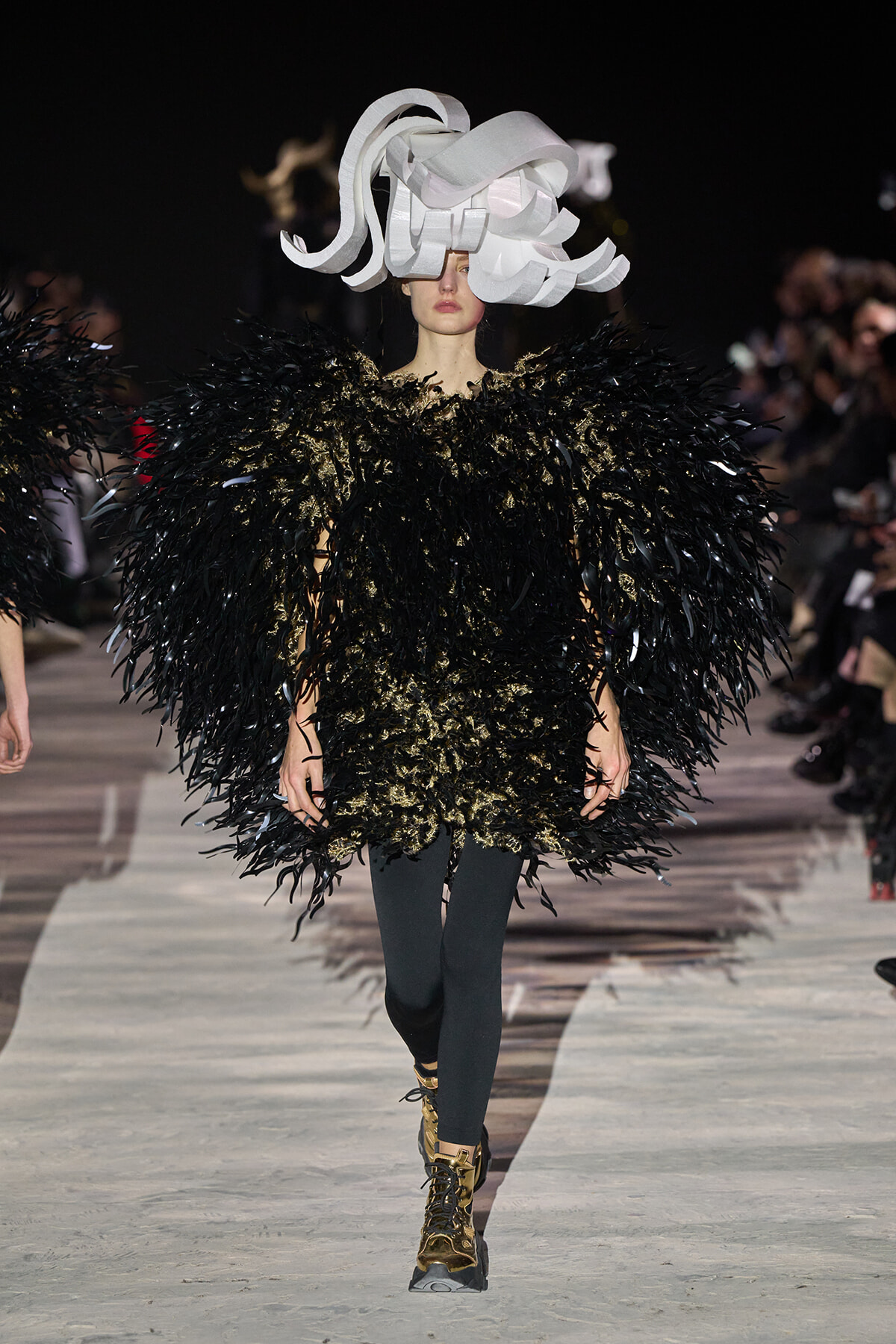 Runway model in a velvety black feathered cape with gold accents and a large white sculptural headpiece, walking the runway.