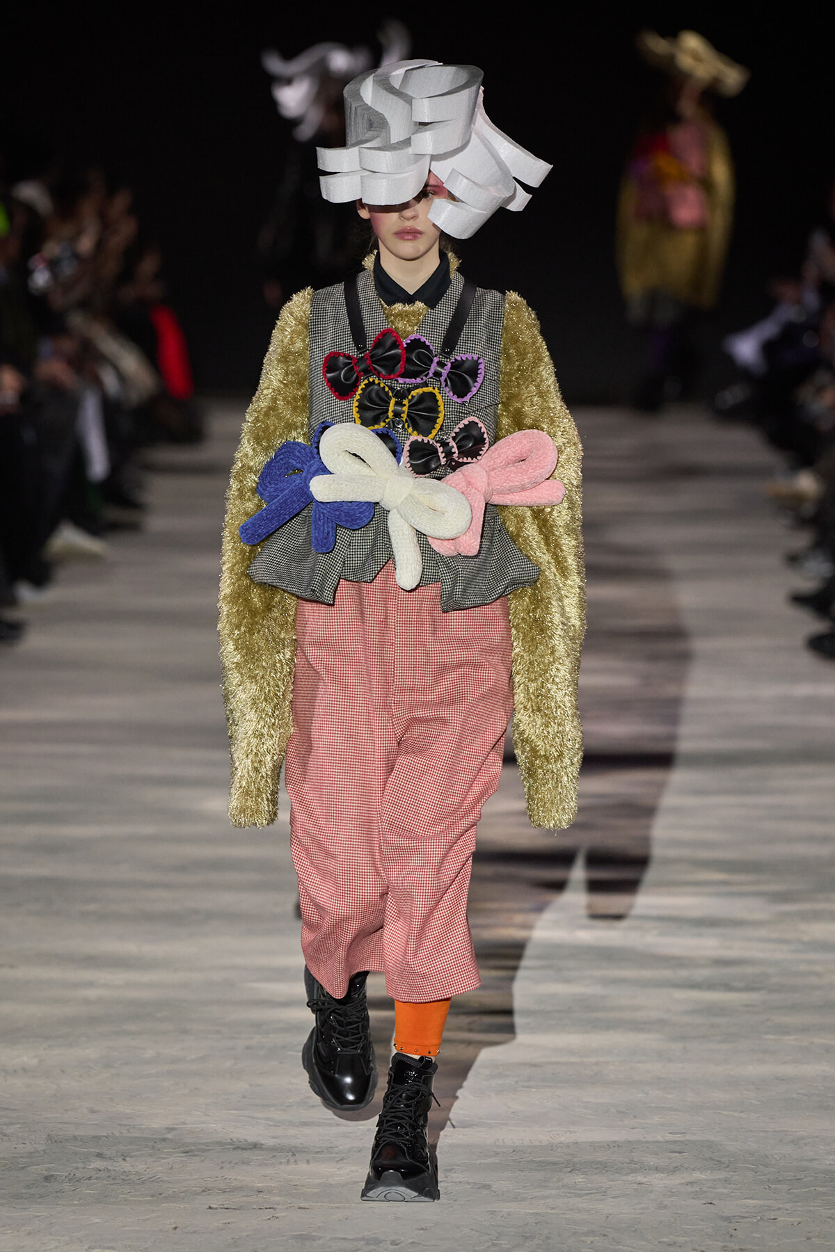 Model on a runway wearing an avant-garde ensemble: large white sculptural hat, gold fuzzy cardigan, pink gingham pants, and colorful bow appliqués.
