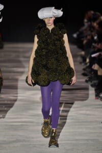 Model walks the runway in a voluminous black-and-gold textured dress, purple tights, gold boots, and a large white sculptural hat.