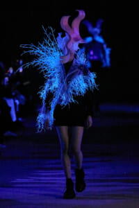 Performer walking in a dark setting, wearing a black outfit with electric blue fiber fringe and a large pink sculptural headpiece under blue lights.