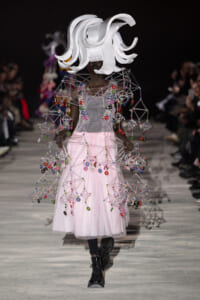 Model on a runway in a pale pink tulle gown with a geometric beaded wire cape and a large white sculptural headpiece.