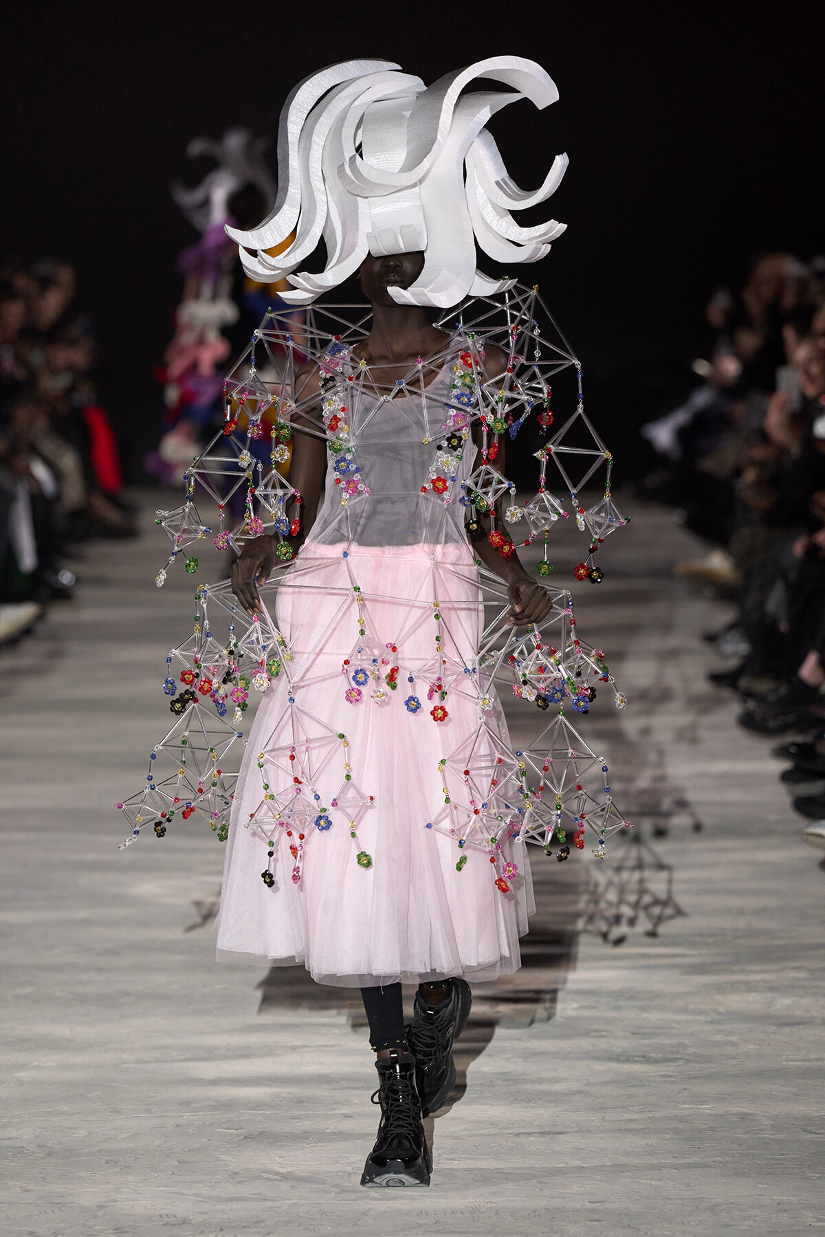 Model on a runway in a pale pink tulle gown with a geometric beaded wire cape and a large white sculptural headpiece.