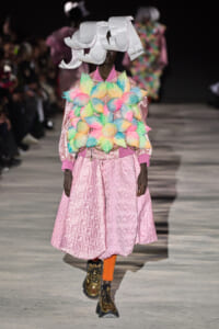 Runway model in a pink quilted dress with a bright multicolored fur bib and a large white paper headpiece, wearing gold boots.