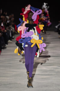 Model on a runway wearing a sculptural outfit made of multicolored knitted bow shapes over a black top and purple pants, with metallic boots.