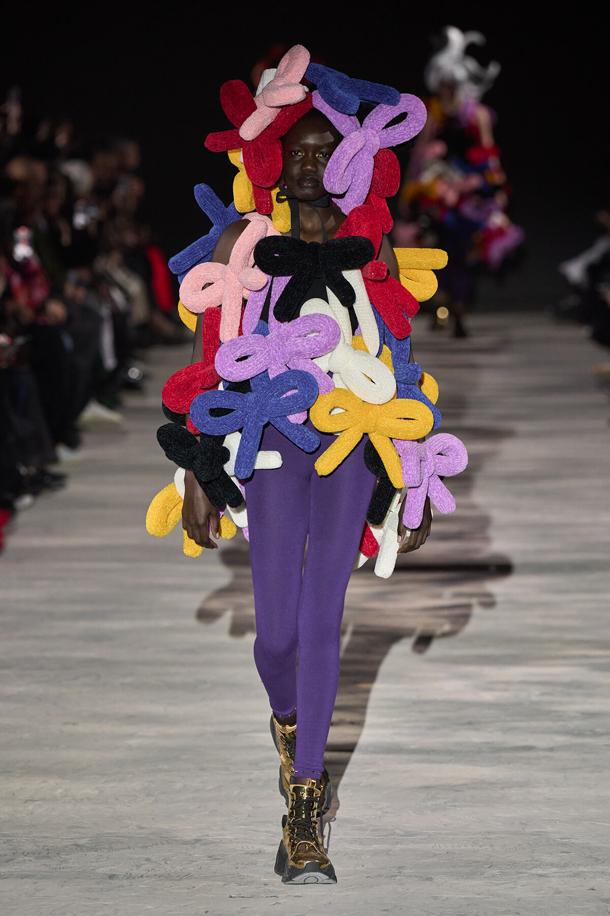 Model on a runway wearing a sculptural outfit made of multicolored knitted bow shapes over a black top and purple pants, with metallic boots.