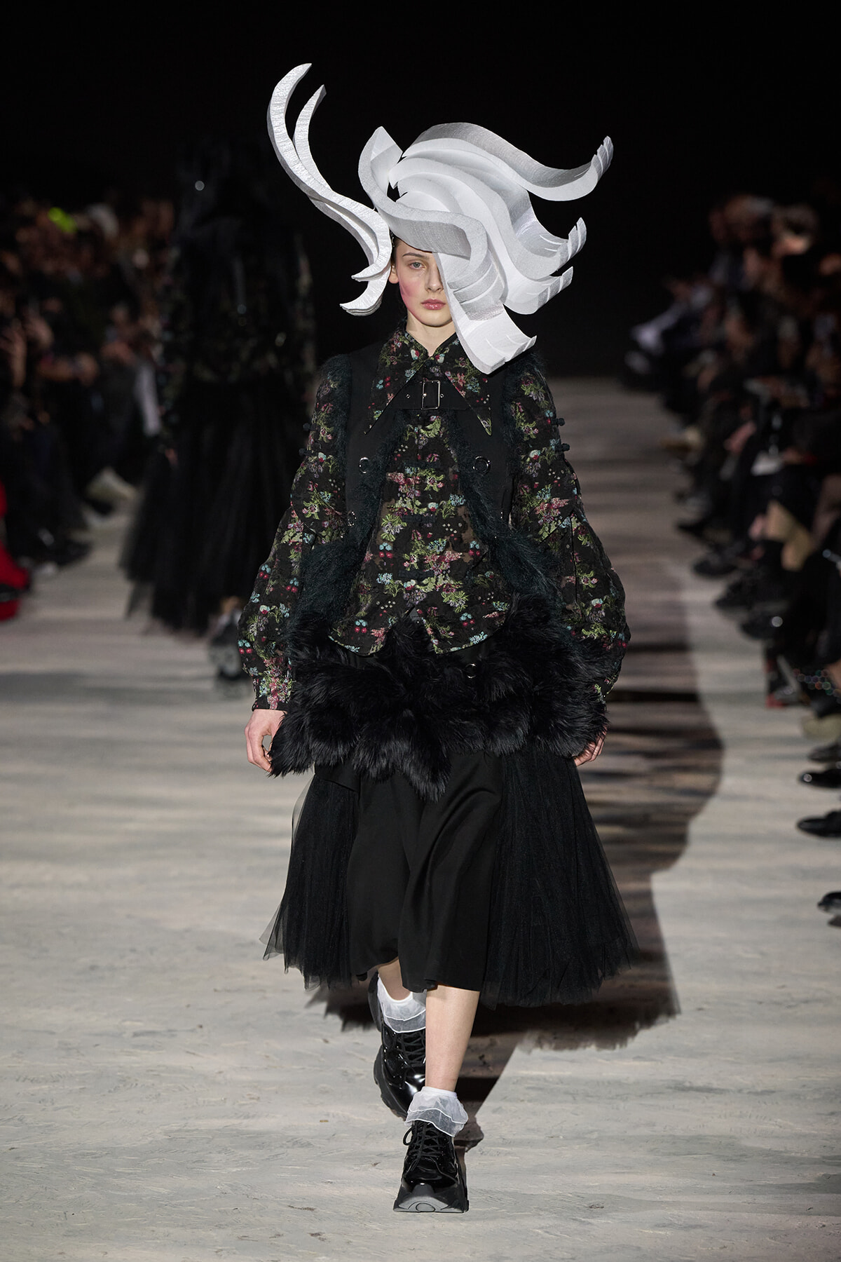 Runway model wearing a large sculptural white headpiece with a dark floral blouse, black fur-trimmed vest, and a black tulle skirt.